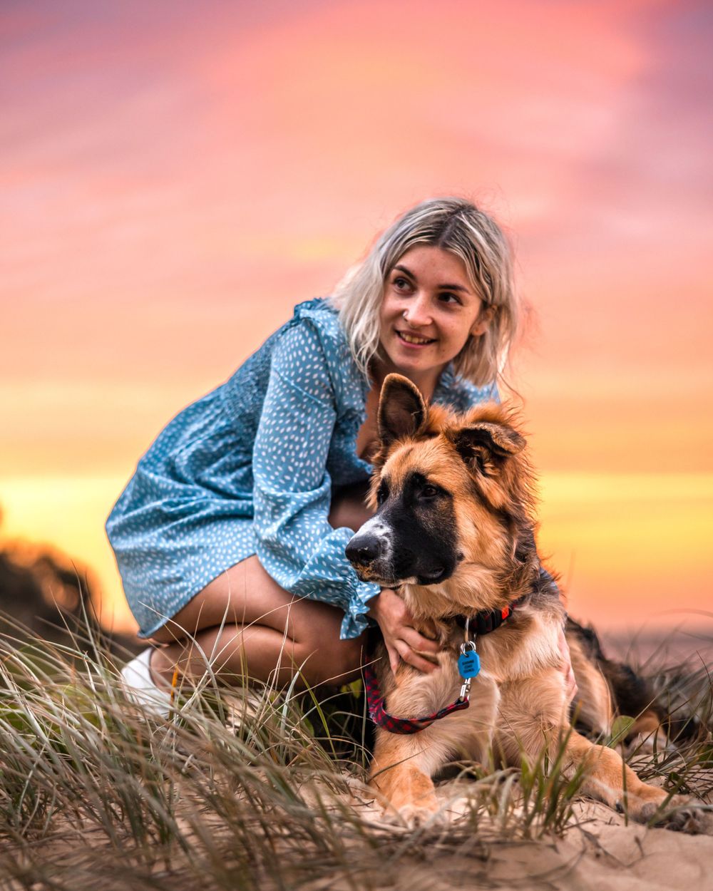 A gorgeous looking female in a vibrant blue dress cuddling her German shepherd dog against AE beautiful soft sunset background