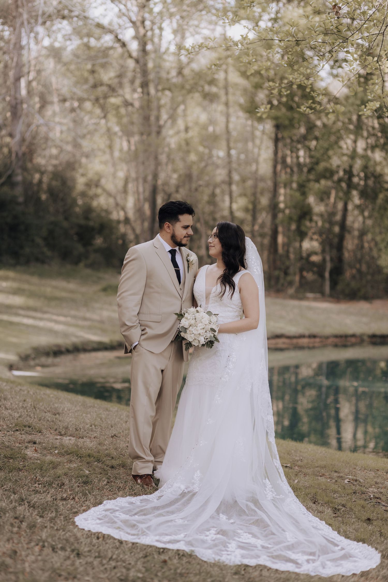 Newlyweds in beige suit and white gown pose by pond during outdoor wedding.