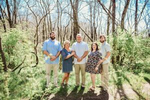Family members laugh and embrace while standing together in a wooded outdoor setting.