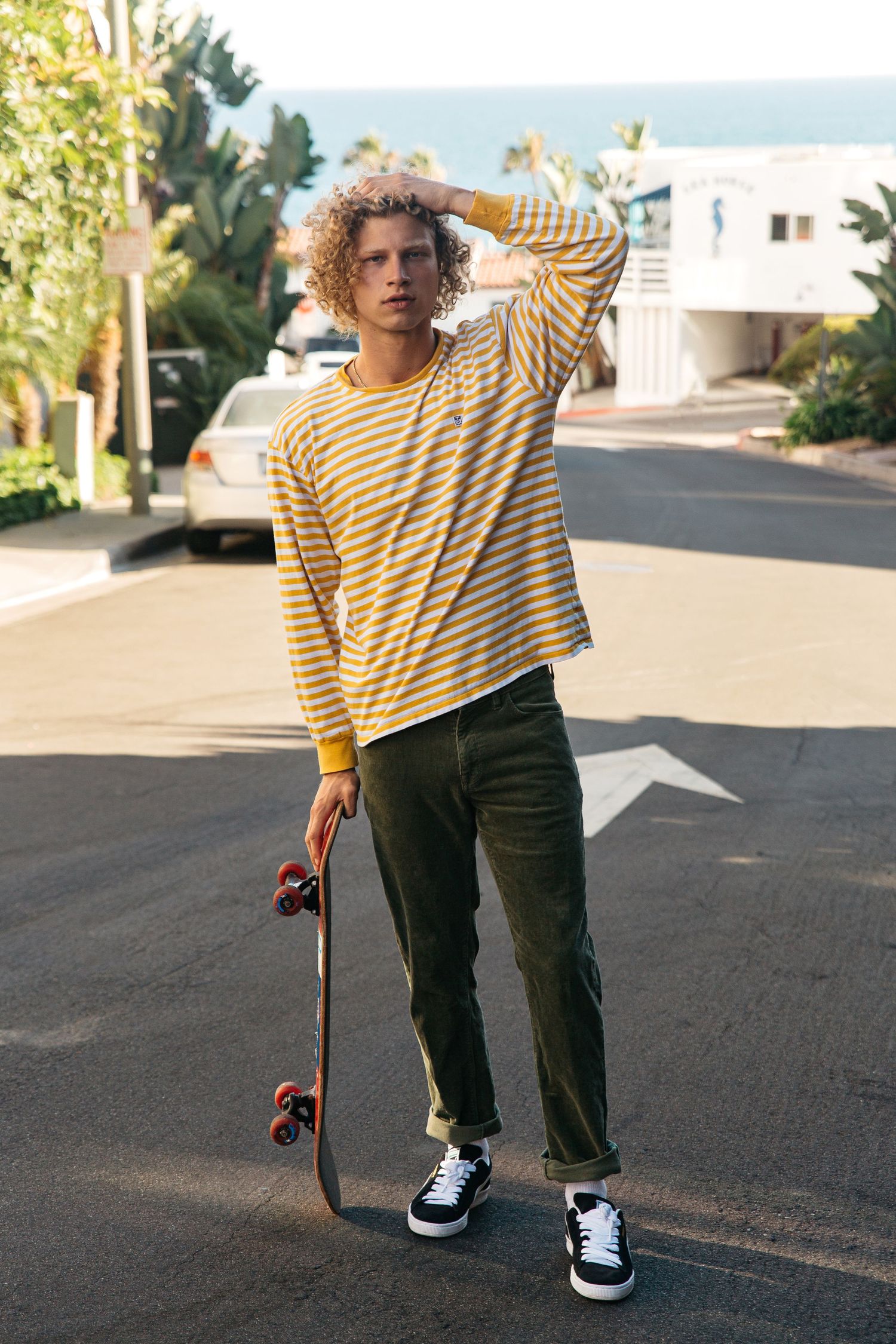 Curly blonde male model holding skateboard on beach town street, San Clemente beach fashion editorial