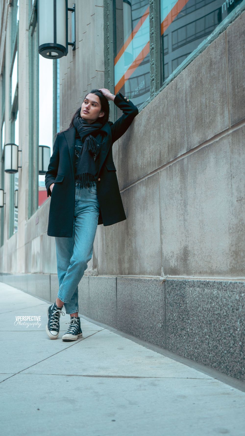 Street style photo of person in black coat and jeans leaning against urban wall.