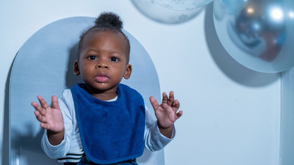 Baby in blue bib raising hands up against light blue background.