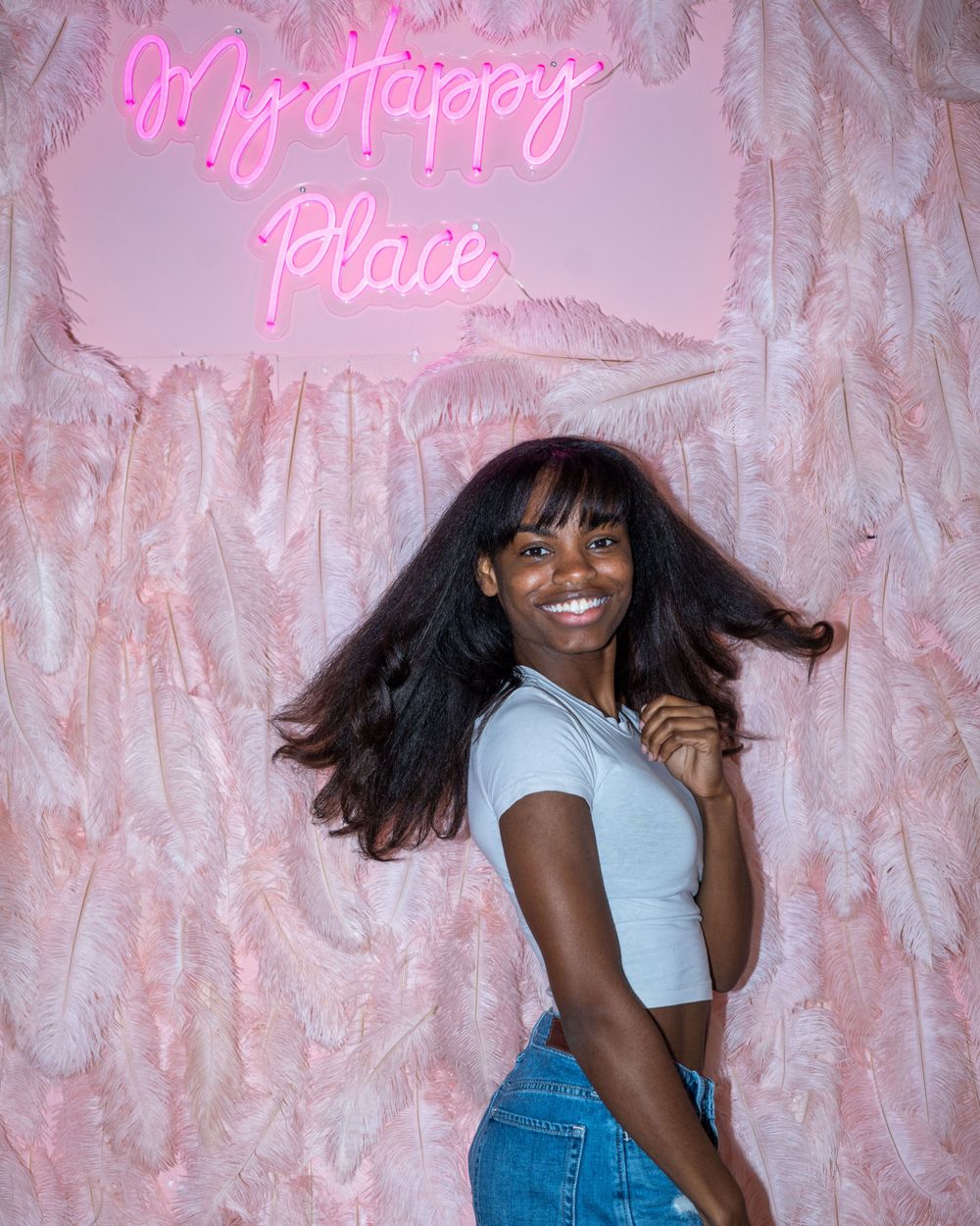 Person posing under pink neon 'My Happy Place' sign against textured backdrop.