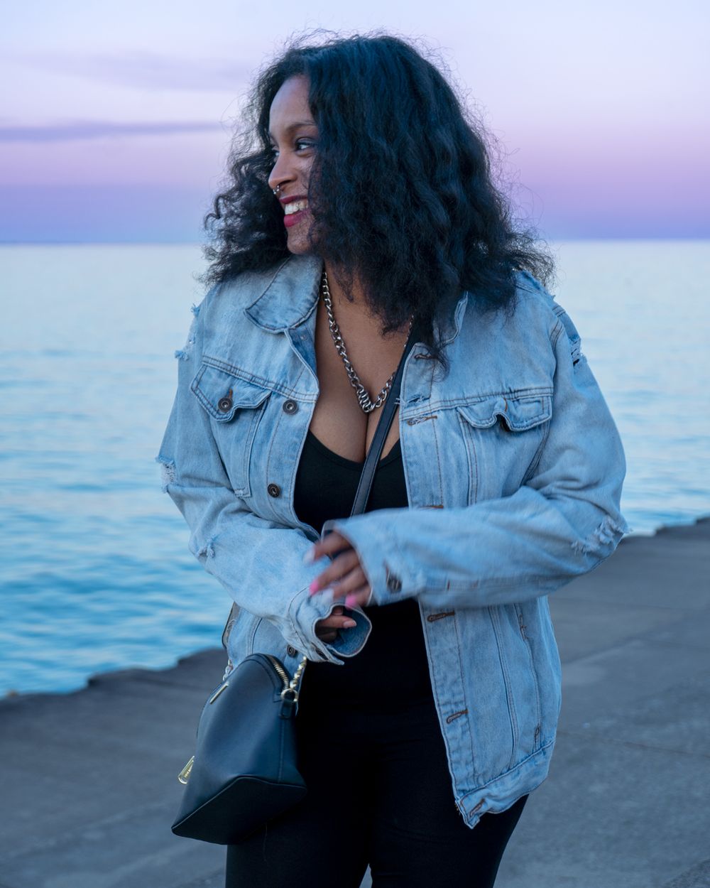 Person in denim jacket smiling by the ocean during sunset.