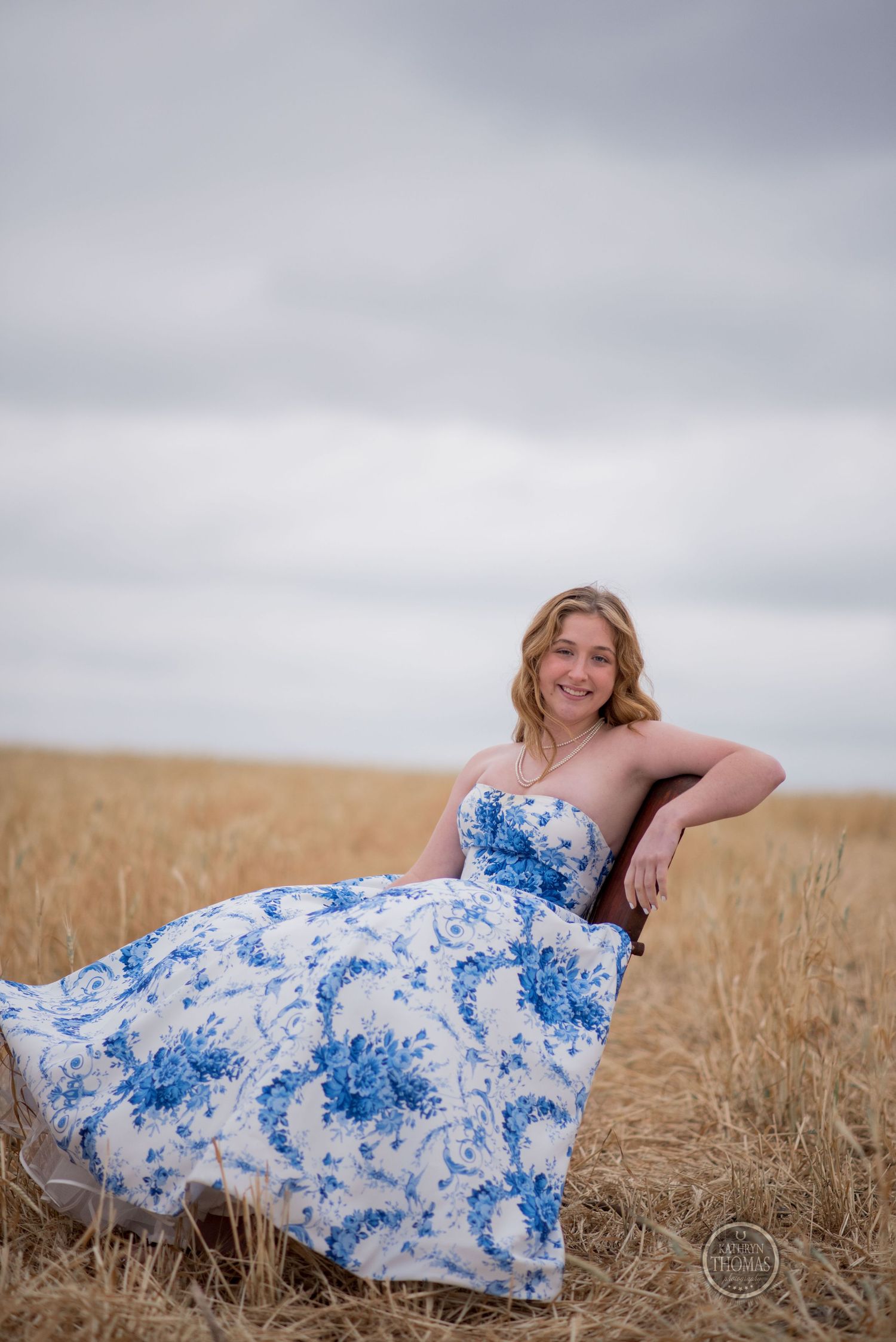 Relaxed portrait in strapless blue floral gown against moody sky and wheat field backdrop.