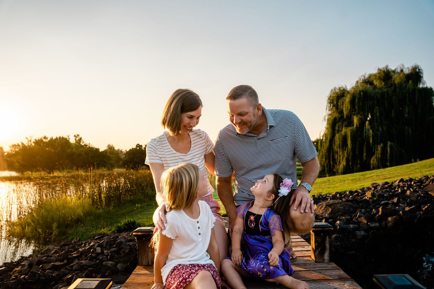 Happy family of four sitting together on dock at golden sunset near peaceful waterway.