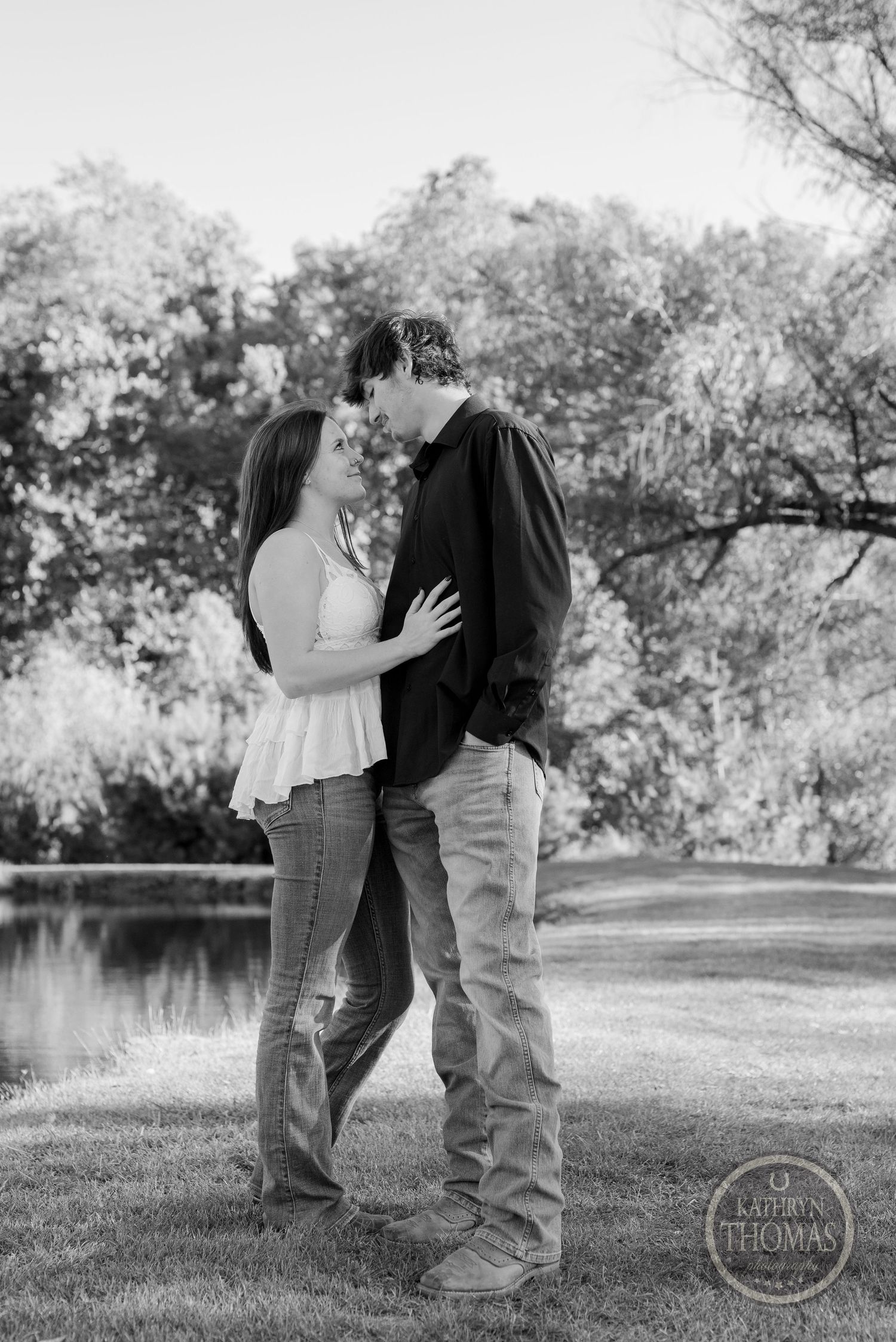 A black and white romantic portrait of a couple by a pond with trees in the background.