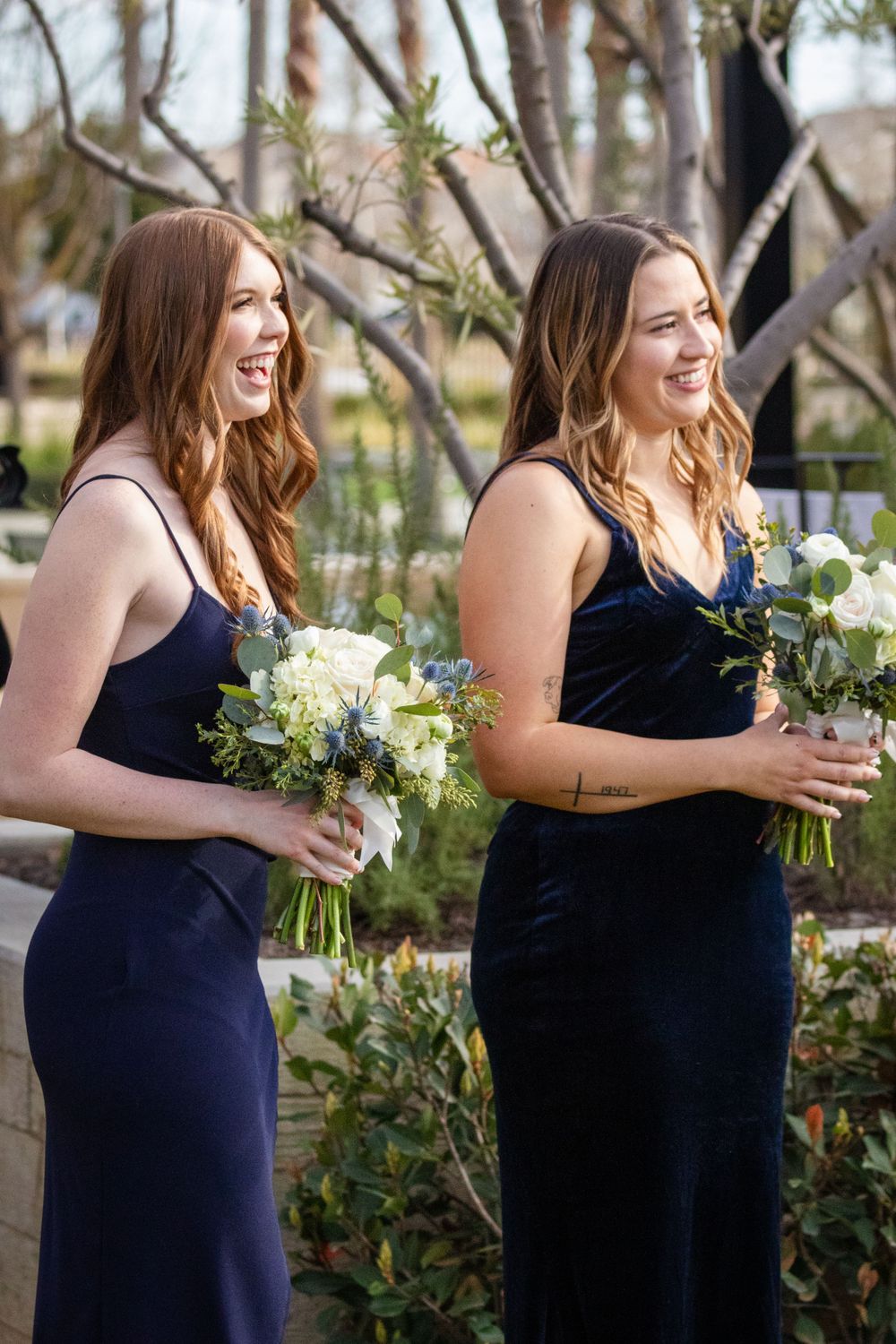 Bridesmaid in navy dress holding white floral bouquet smiles during outdoor wedding ceremony.
