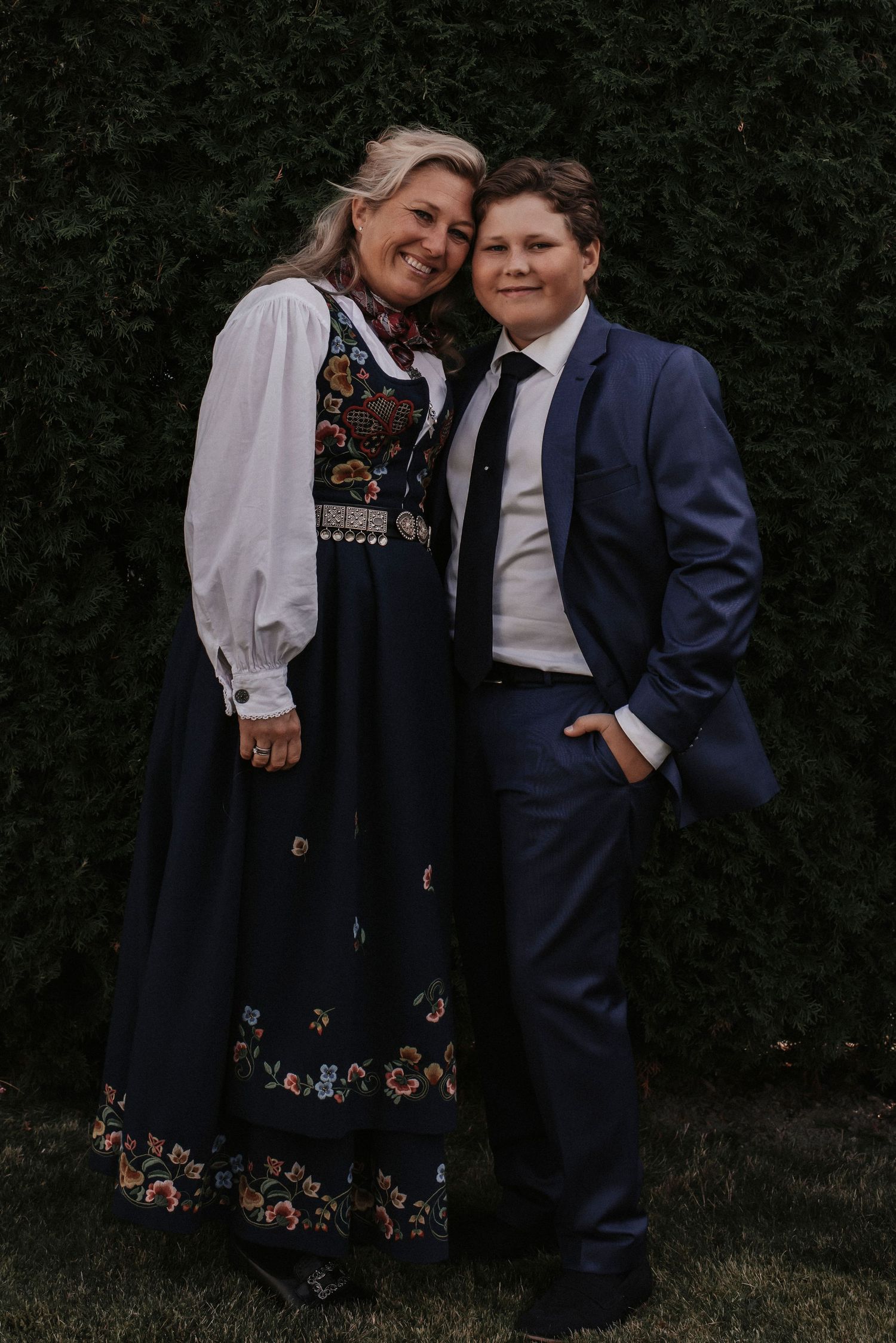 A pair wearing formal attire with one in a traditional Norwegian bunad dress pose against a hedge.