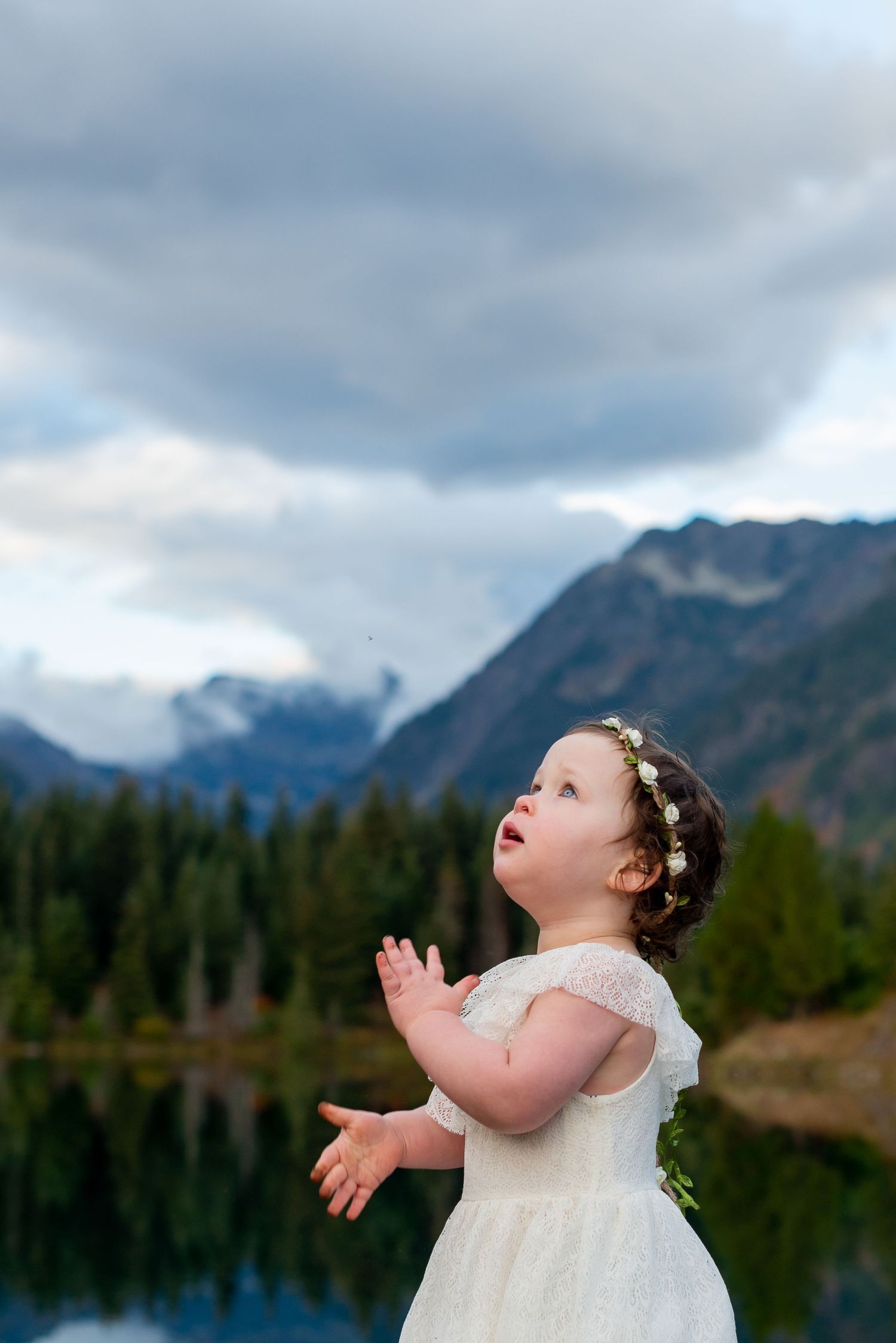 Child in white dress poses against mountain landscape with dramatic cloudy sky.