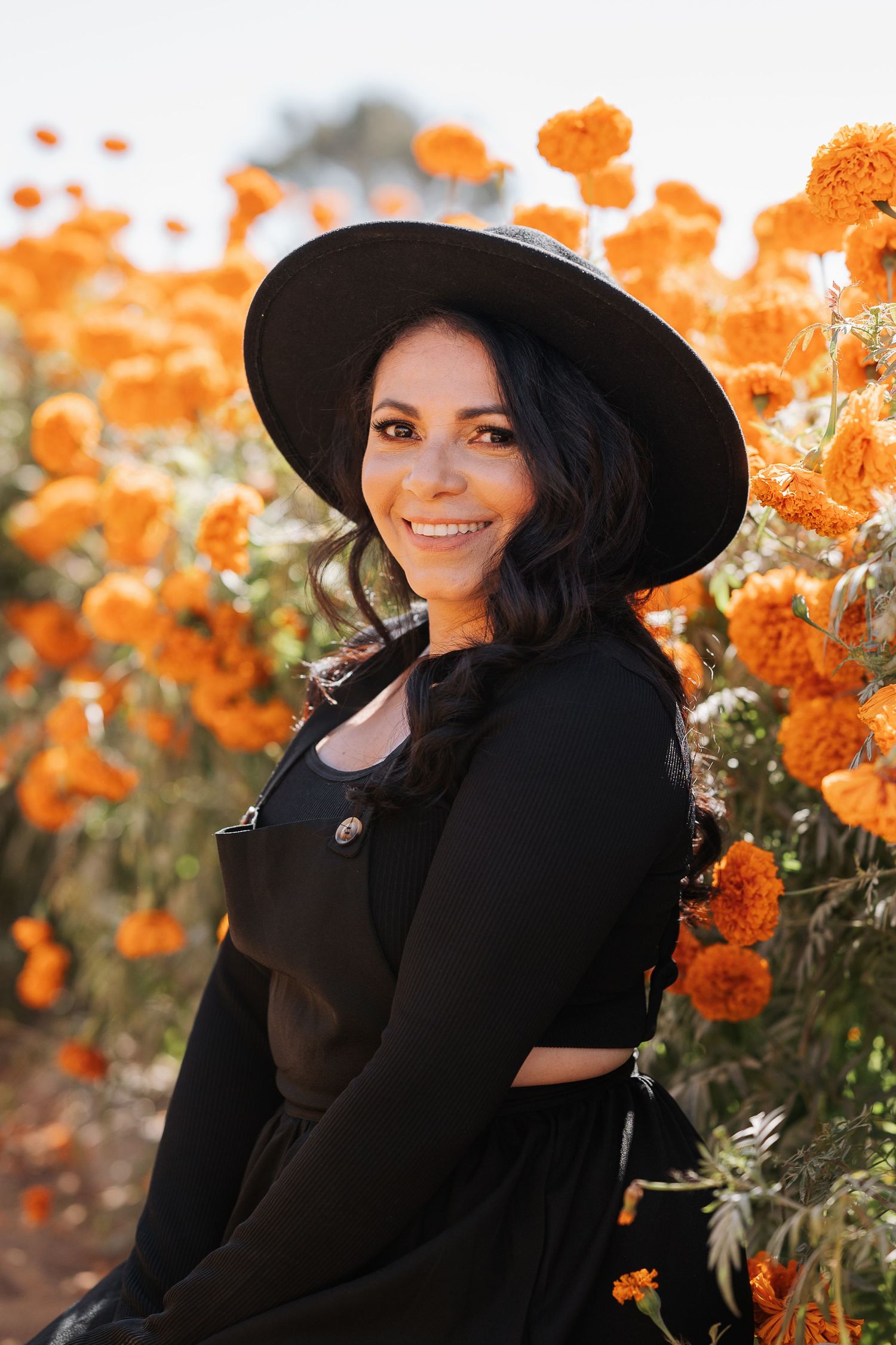 Portrait in a black dress and hat among vibrant orange flowers in natural light.
