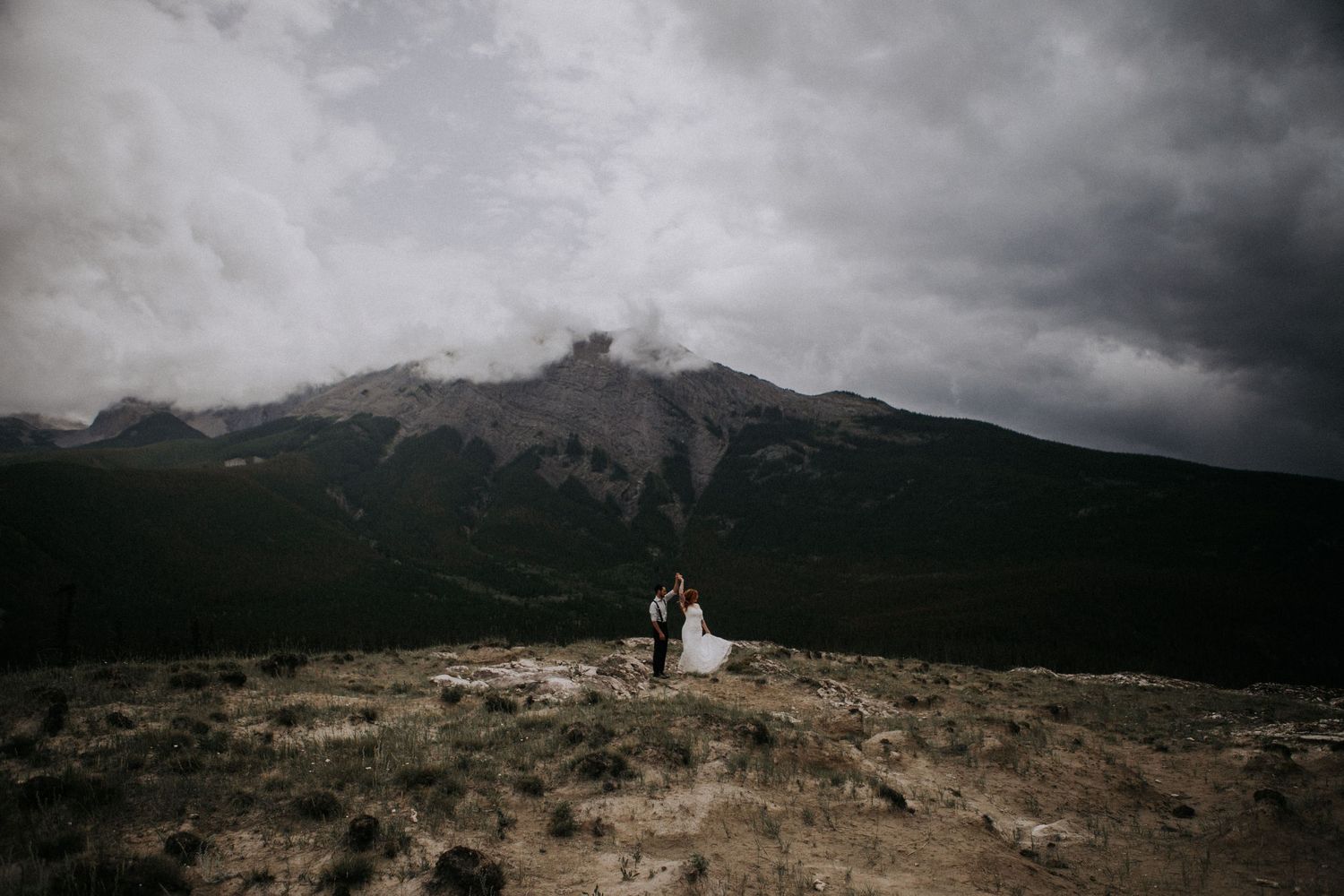 A tiny couple stands silhouetted against dramatic mountain peaks and moody storm clouds on a barren hillside.