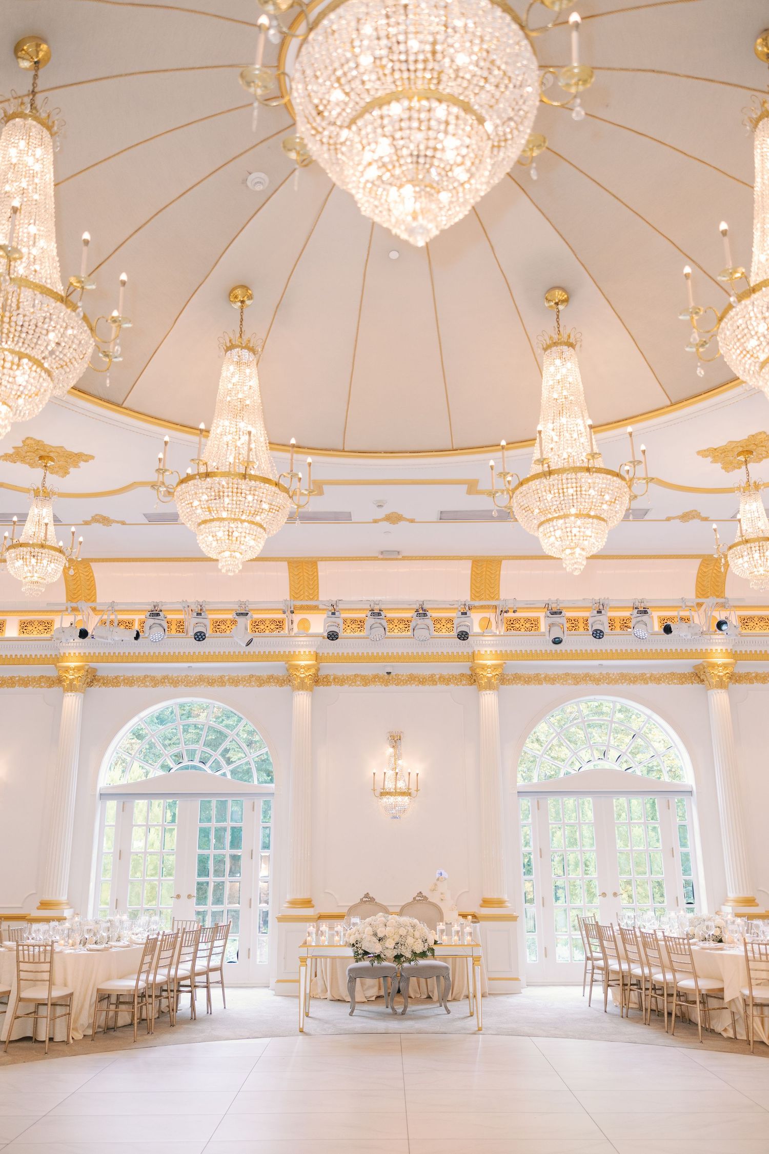 Elegant gold and white ballroom with crystal chandeliers and arched windows set for wedding reception.