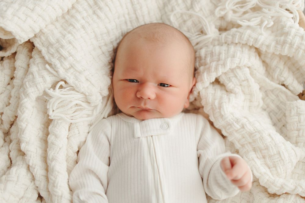 A small baby wearing a white knit outfit lies on soft cream colored blankets.