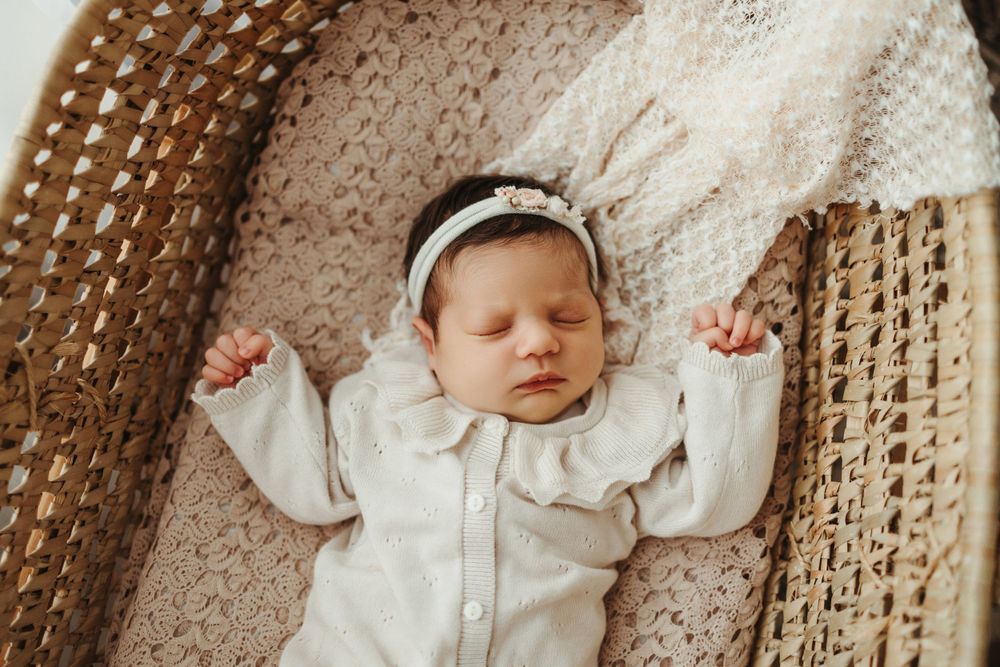 Sleeping baby in white outfit lying on brown knitted blanket with arms outstretched.