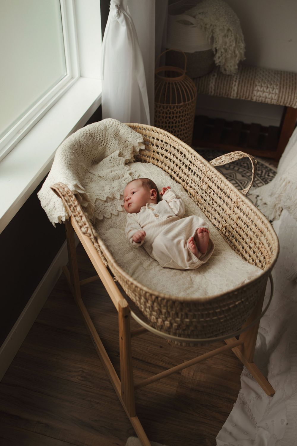 A woven bassinet sits near a window with warm natural light illuminating a white interior and wooden stand.