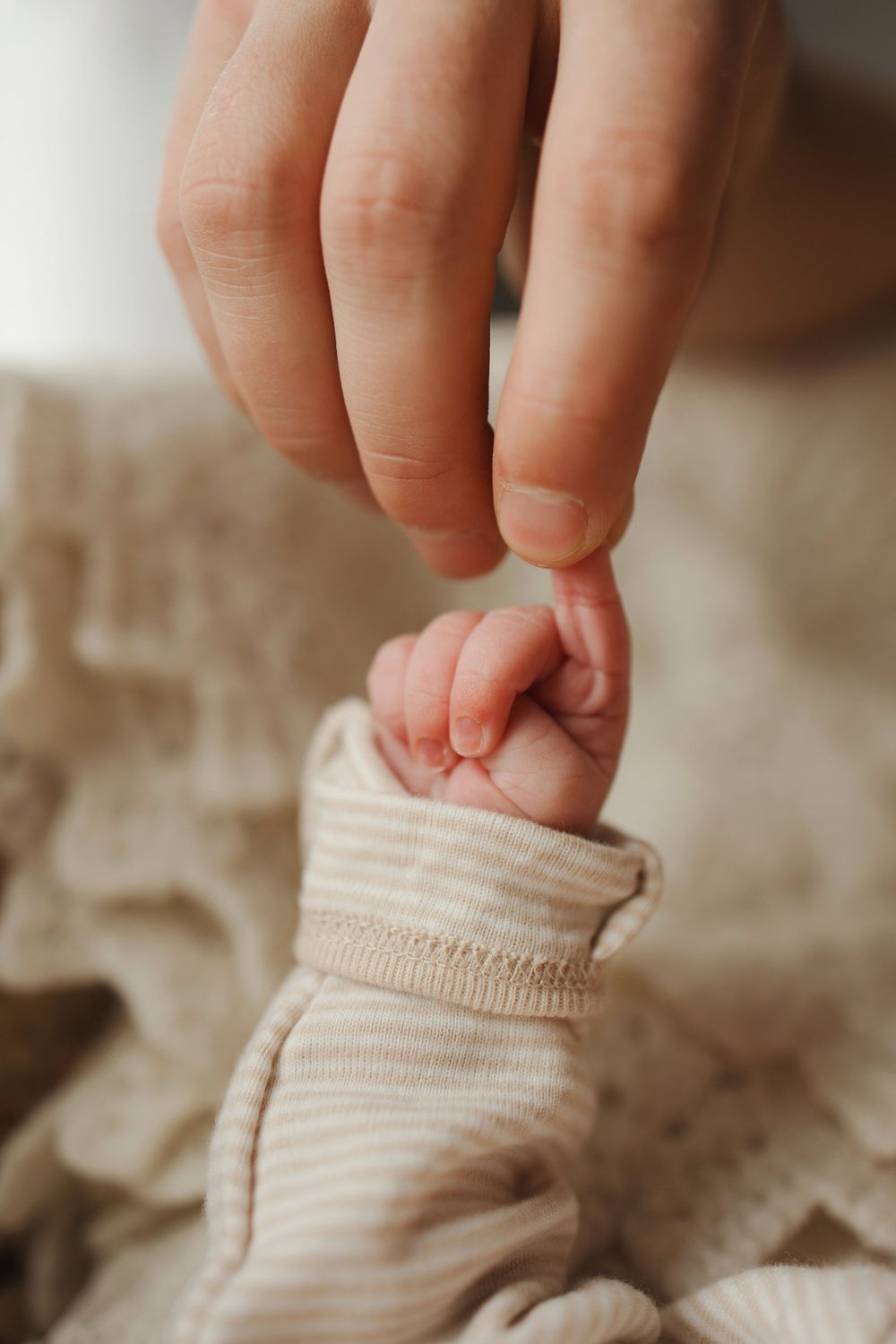 A tender close-up of an adult's finger gently touching a tiny newborn baby's hand in soft beige clothing.