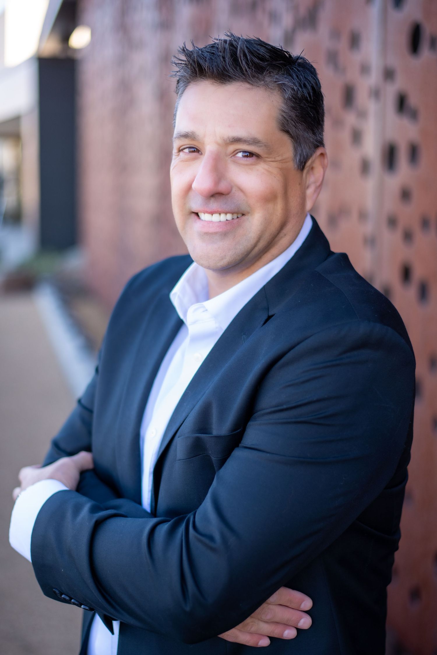 A professional headshot of a man in a dark suit jacket against a brick wall background.