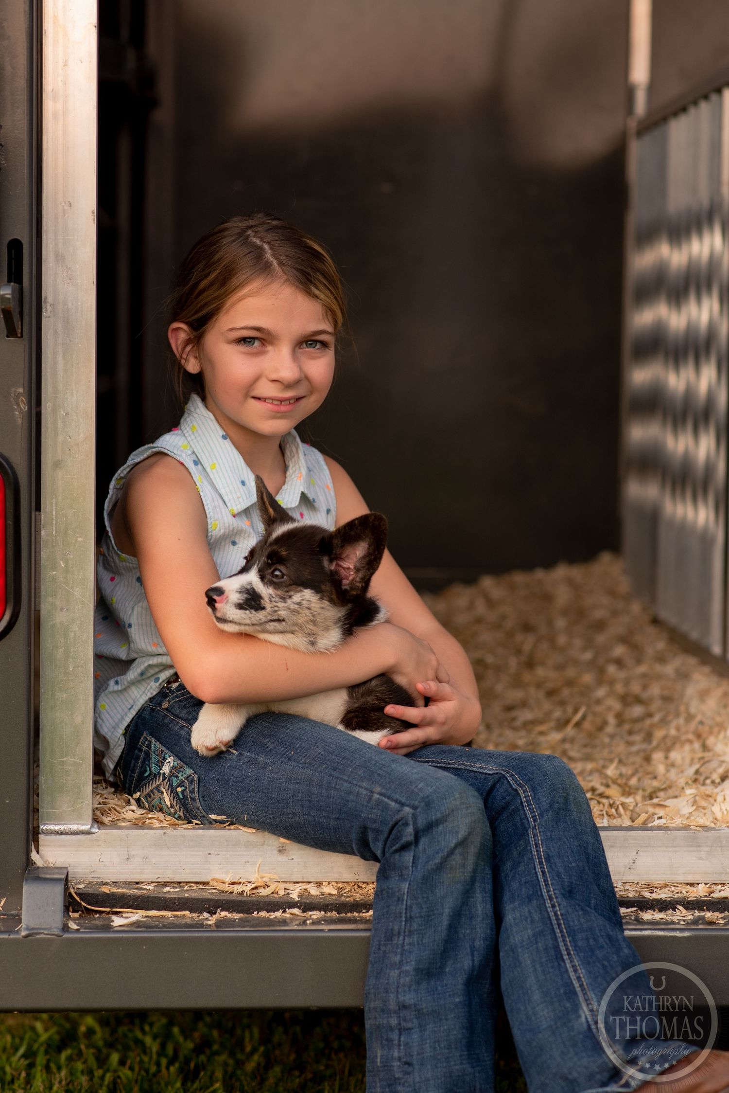 A young person sits in a barn doorway holding a cat while surrounded by straw bedding.