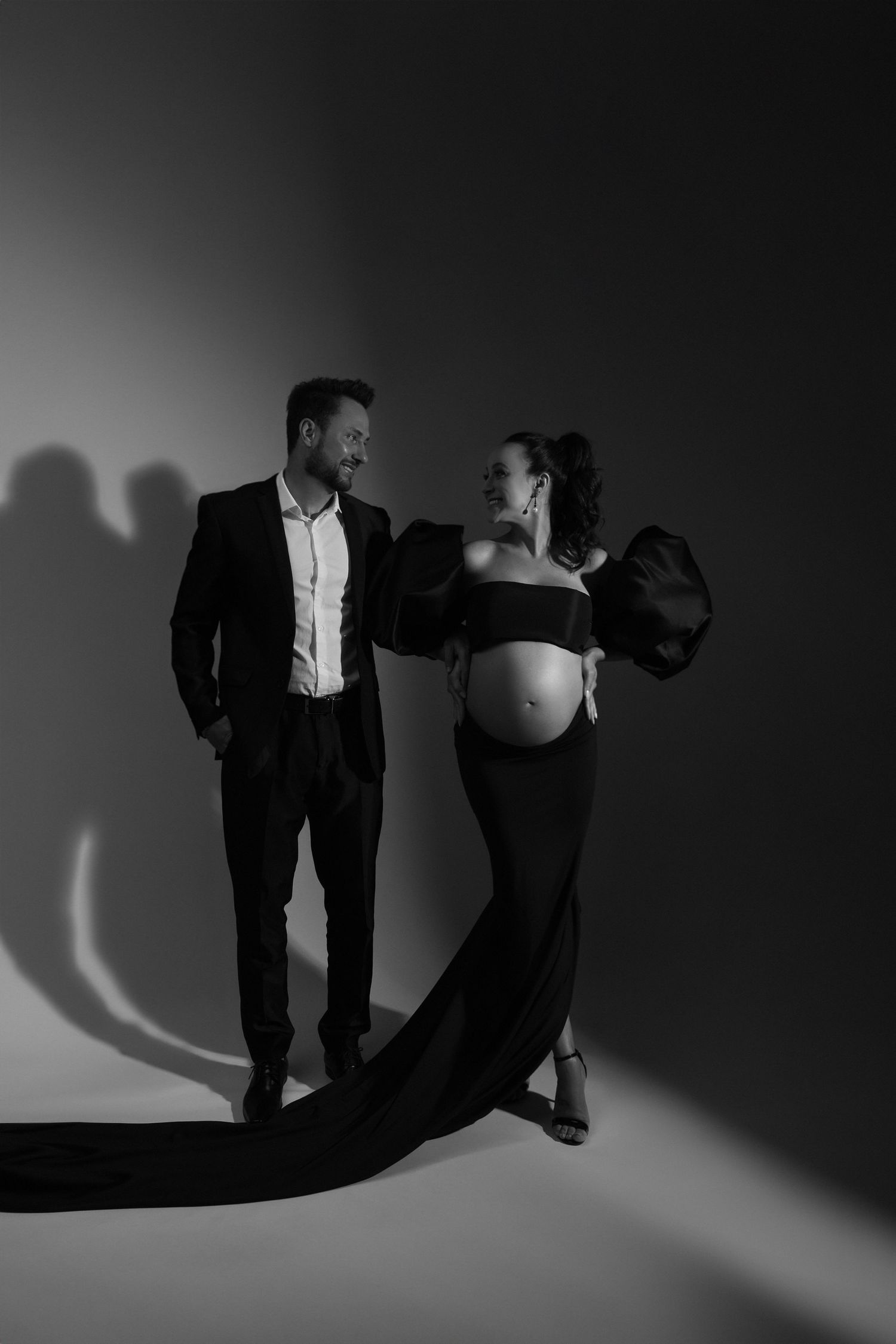 Dramatic black and white maternity photo with flowing dress against dark background.