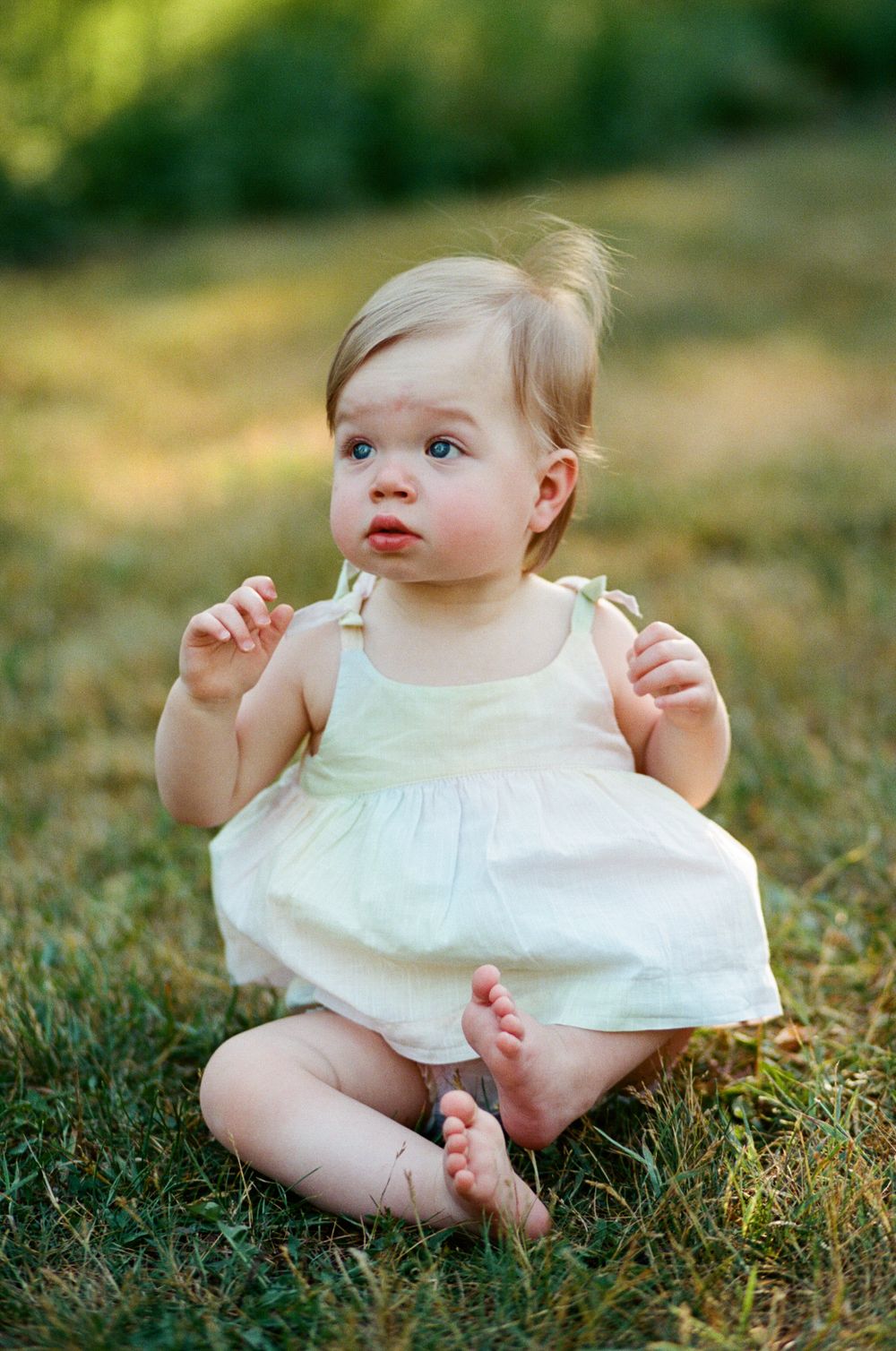Adorable blue-eyed baby girl in white sundress sitting barefoot on green grass outdoors in summer.
