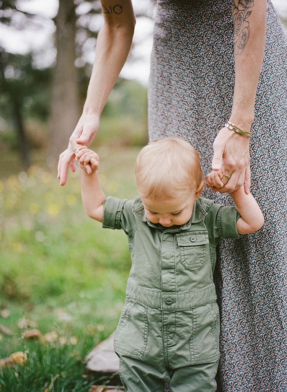 A baby in a green romper learning to walk while holding mother's hands in a park setting.