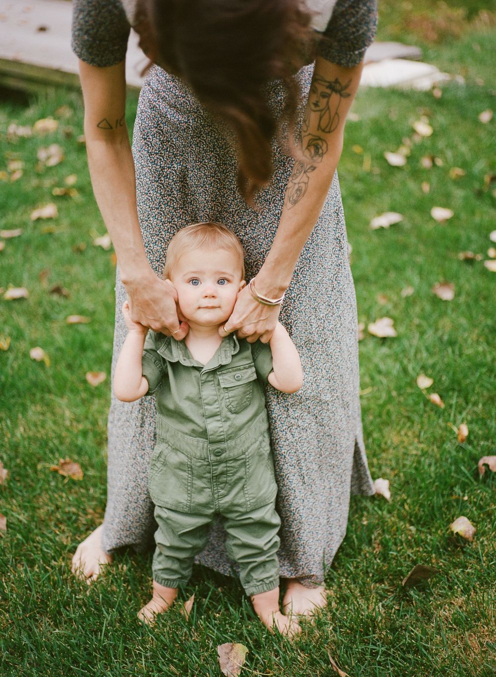 Tattooed mother helping her blue-eyed baby stand on a grass lawn covered with autumn leaves.