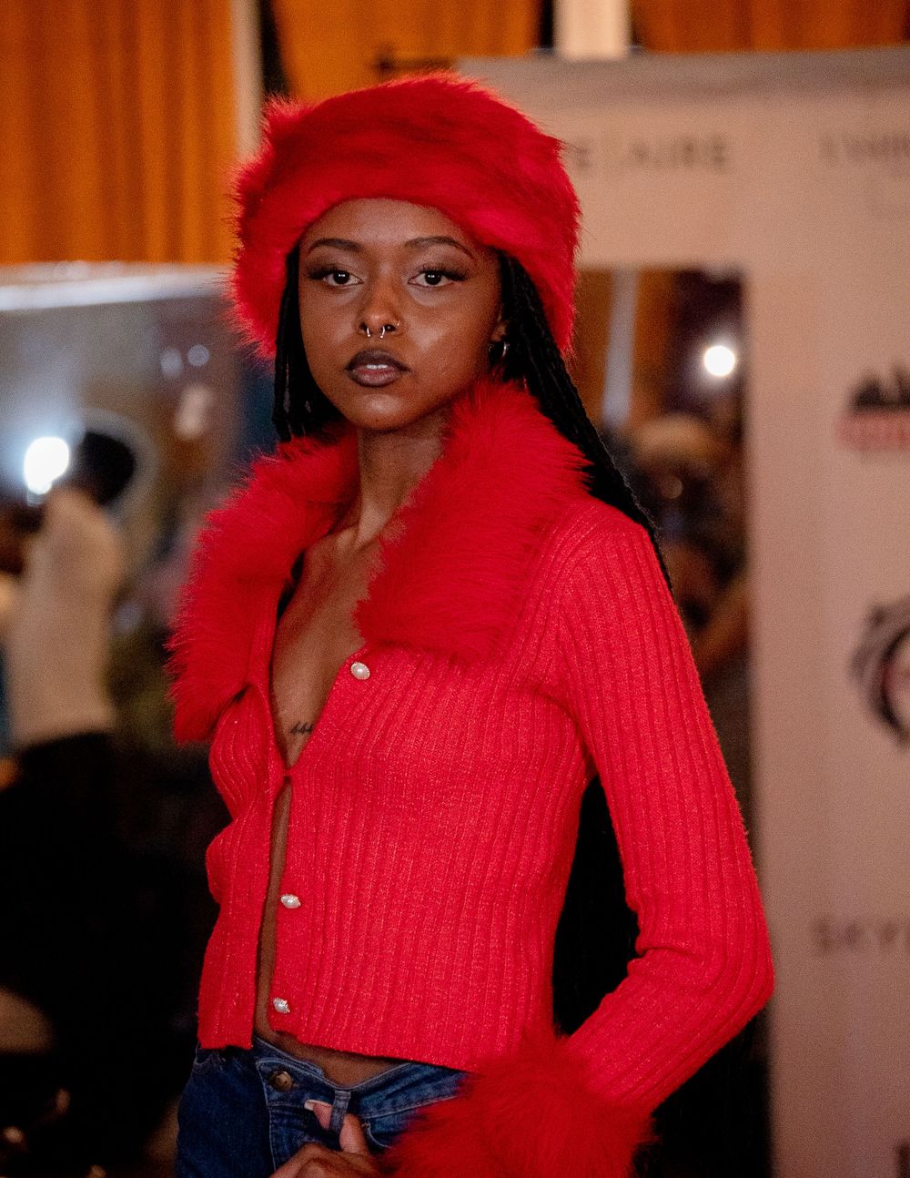 Fashion model in bright red cardigan and matching furry hat poses against event backdrop.