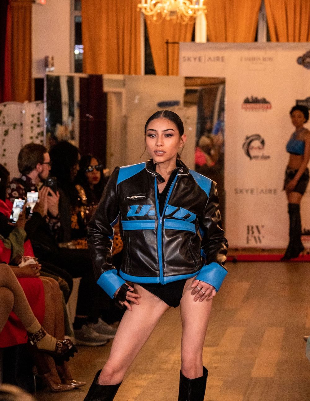 Model walks runway in blue and black racing-inspired jacket during fashion show.