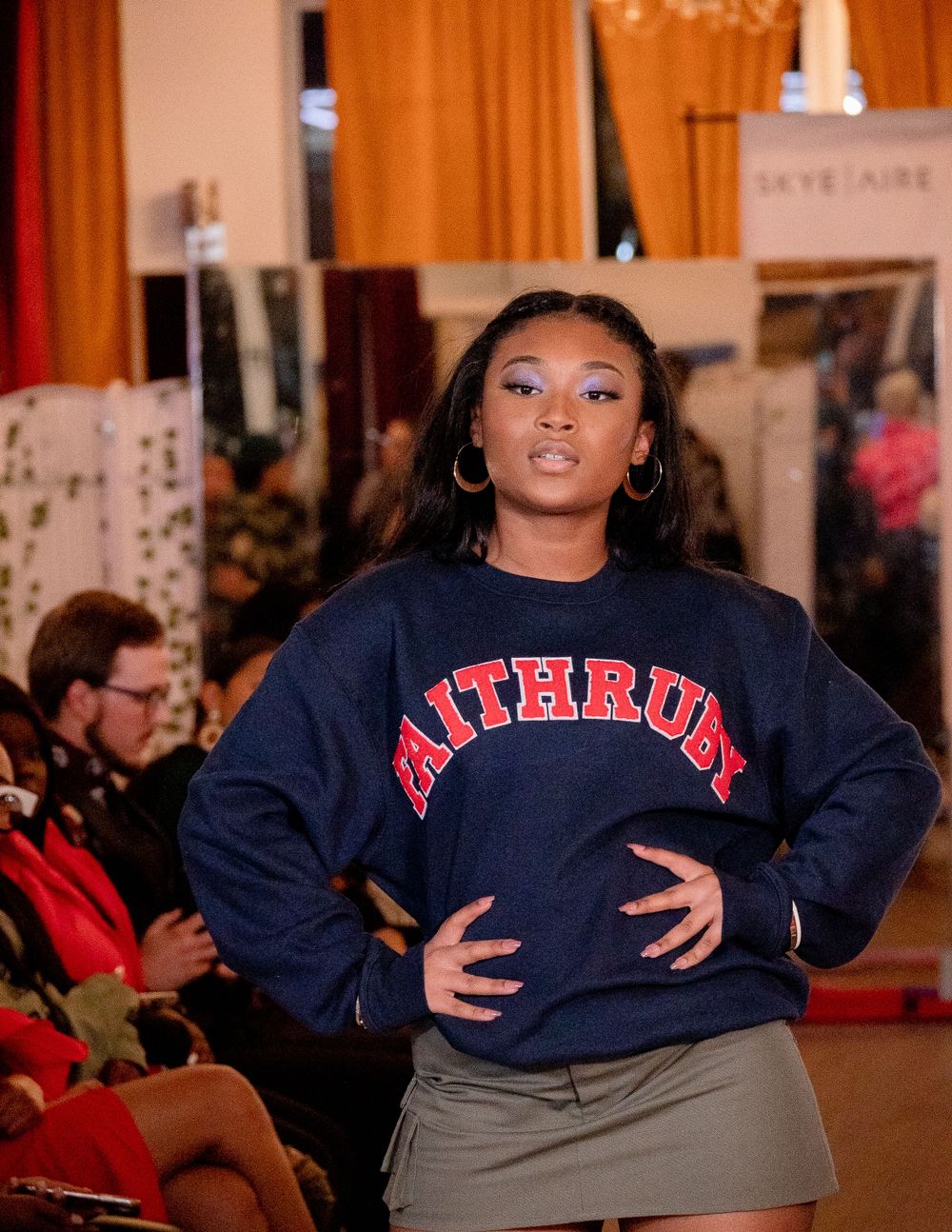 Fashion model in navy blue sweatshirt strikes pose on runway with orange curtains in background.