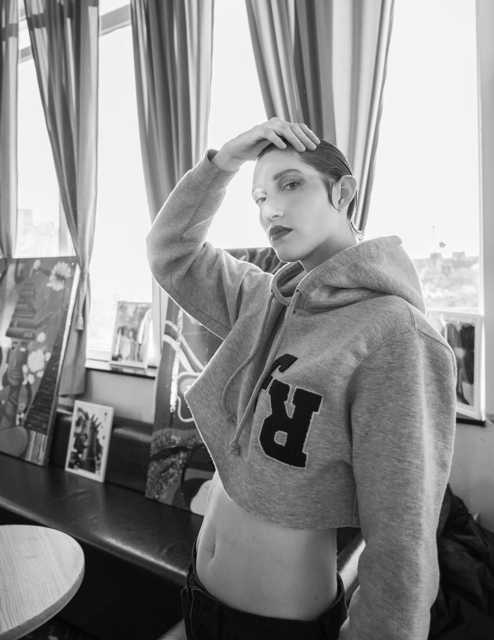 Black and white photo of model in cropped gray hoodie posing by window.