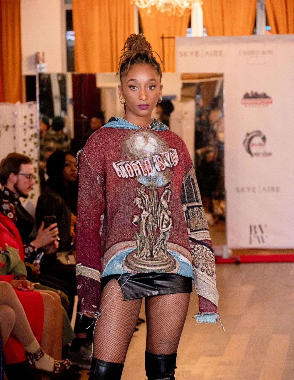 Model wearing burgundy graphic sweater and black shorts walks runway at fashion show with orange curtain backdrop.