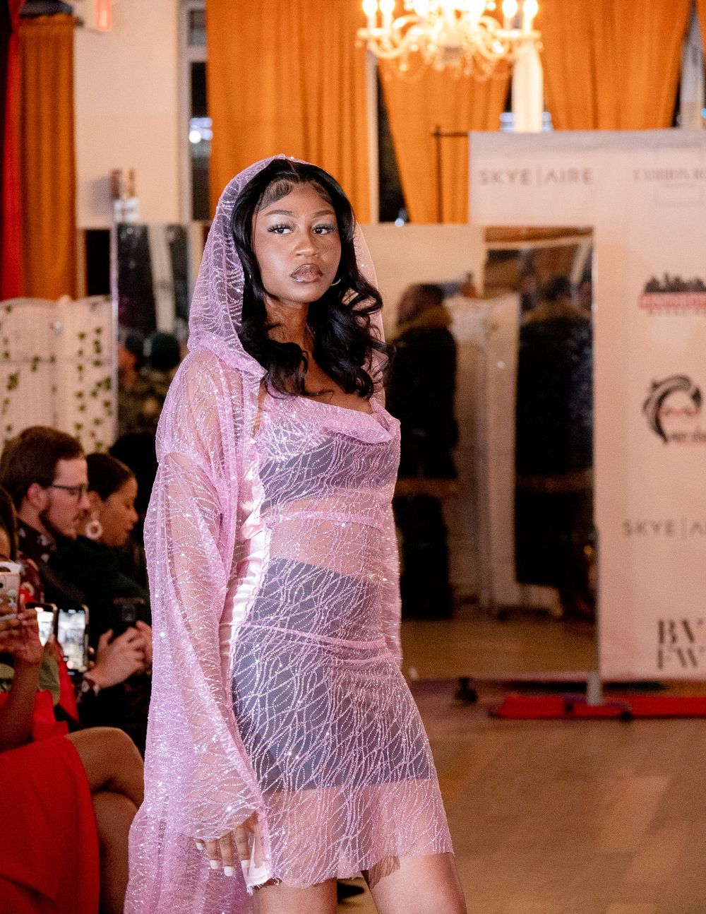 Model in sparkly pink hooded dress poses on runway with chandelier and orange curtains in background.