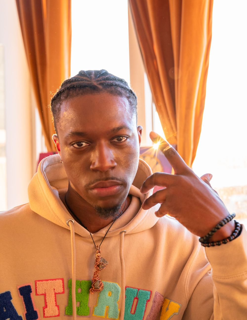Model wearing beige hoodie with colorful lettering poses against orange curtain backdrop.