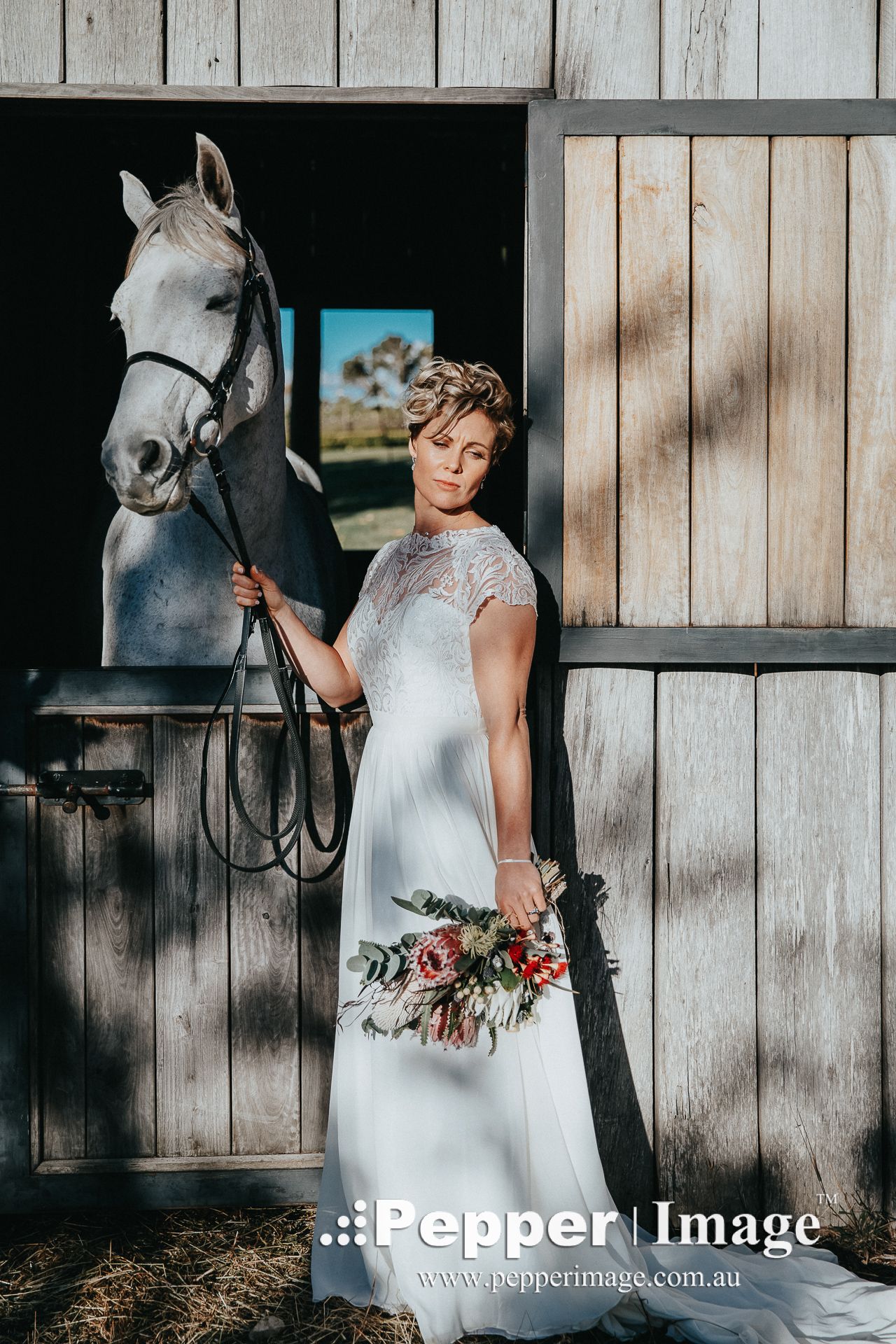 A bride in a white lace gown holds a bouquet while standing beside a white horse at a rustic barn wedding.