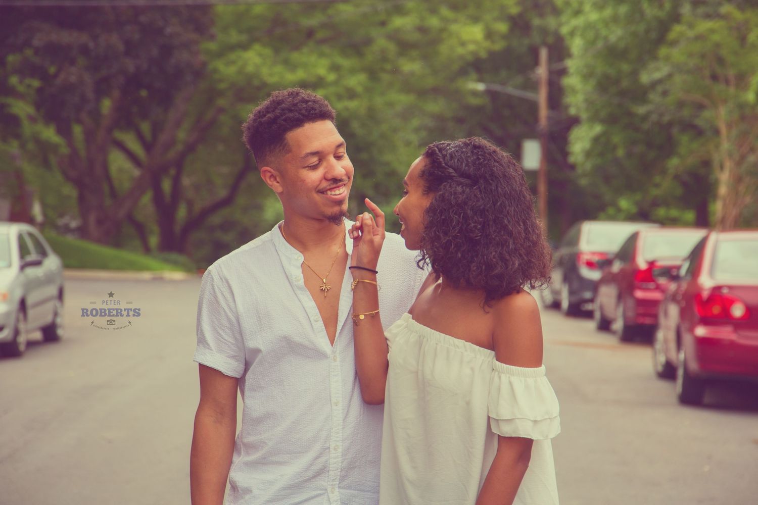 Young couple sharing a tender moment on a tree-lined street in casual white outfits.