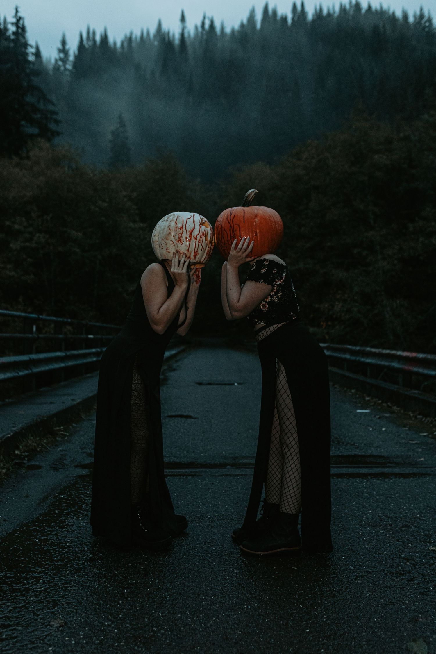 Two figures in black dresses holding pumpkins on foggy mountain road at dusk.