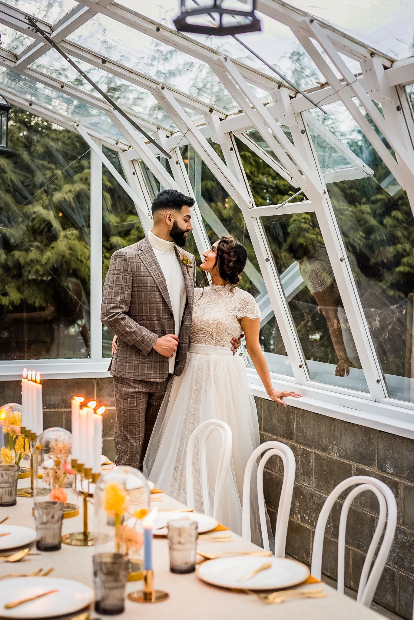 A couple dressed in their wedding attire stands in their venue, a greenhouse, and smile together as they get ready for their reception.