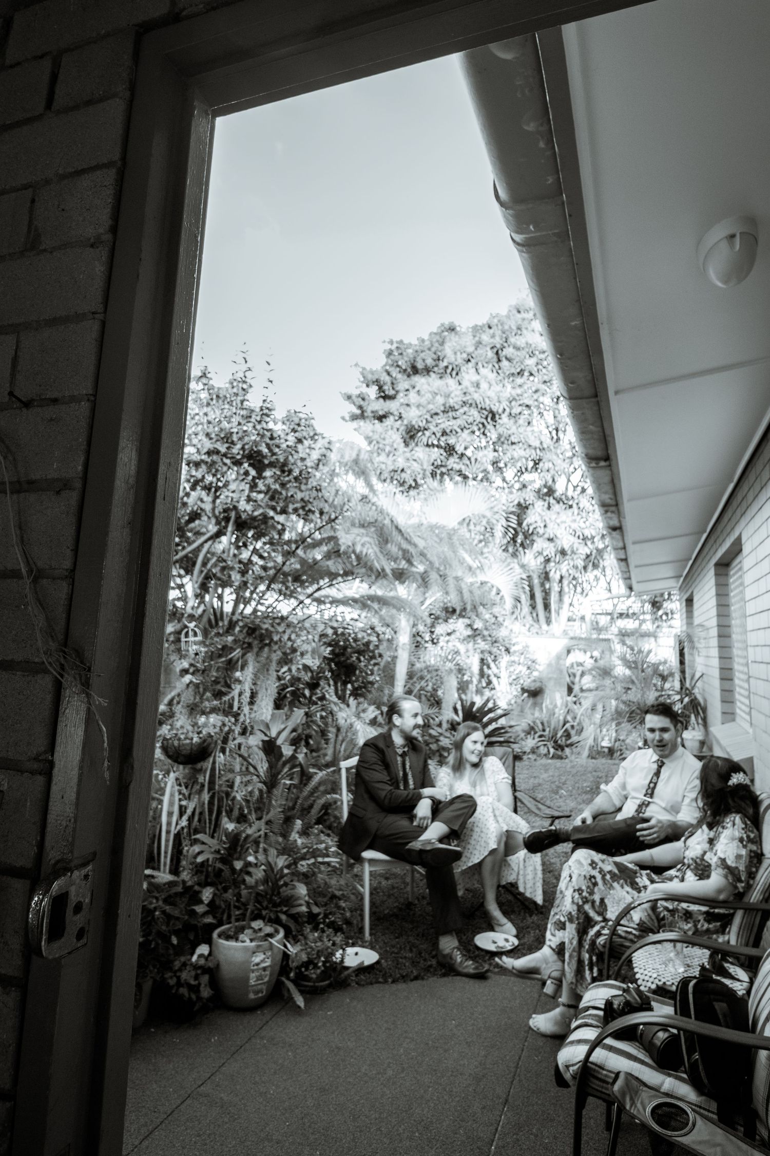 A black and white photo of a group gathering on a covered porch with plants and trees in the background.