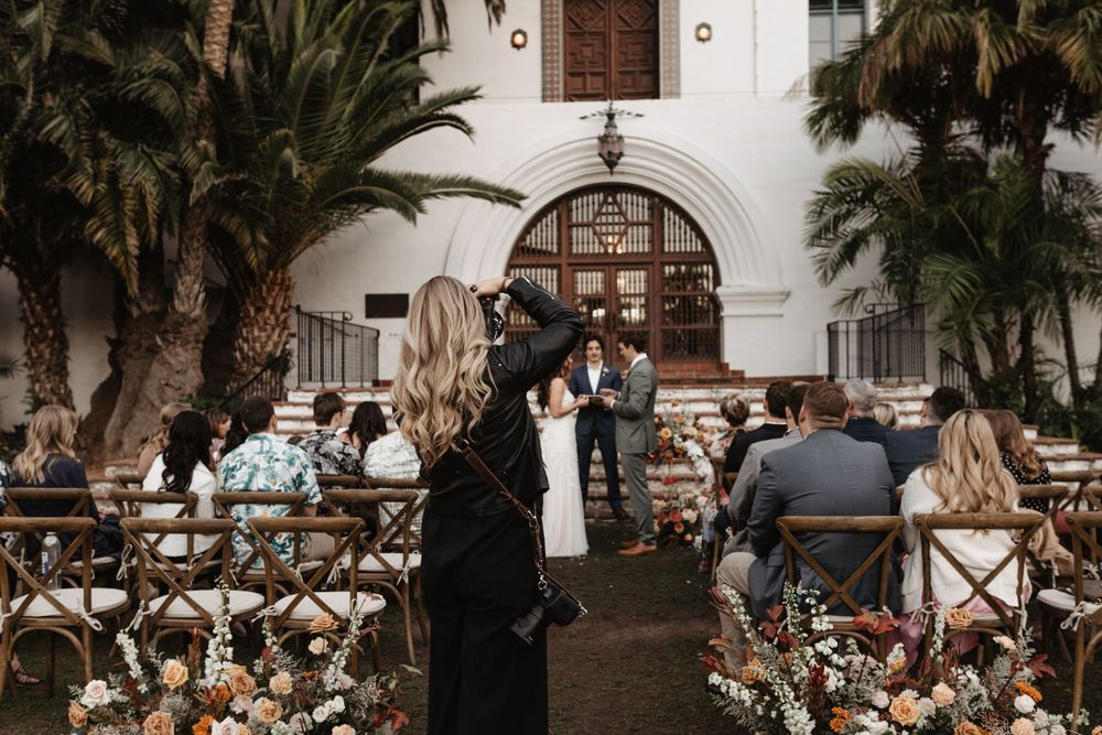 A wedding ceremony takes place at a Spanish-style venue with palm trees and wooden chairs.