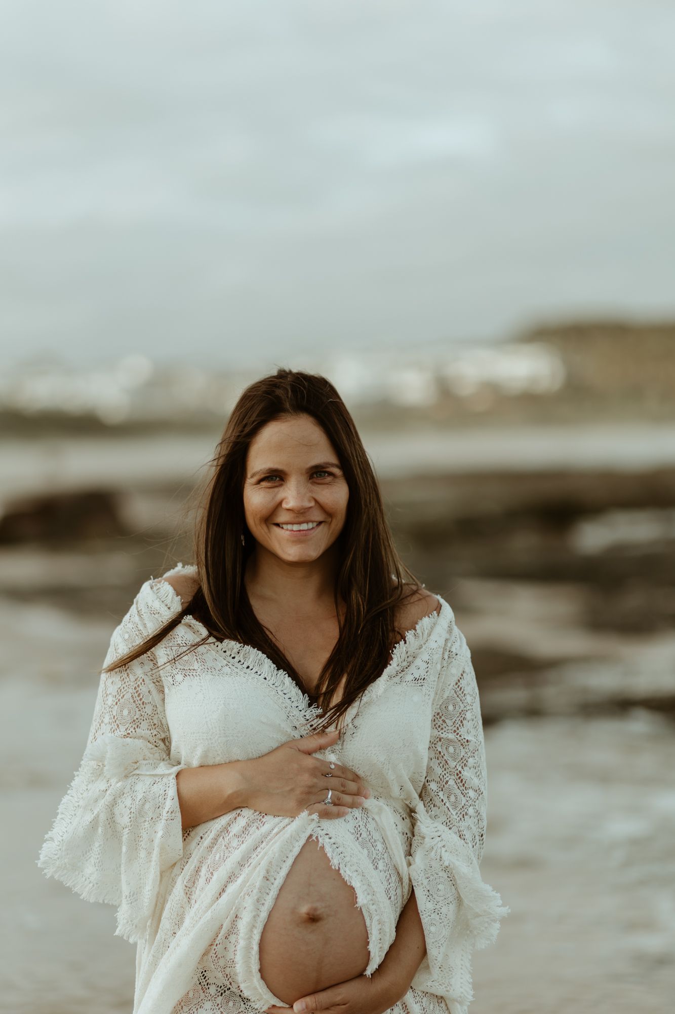 Expectant mother in white lace dress smiles on rocky beach.
