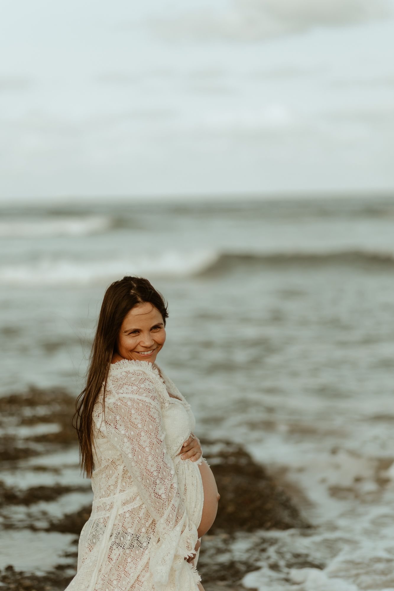 Expectant mother in white lace dress poses on rocky beach at sunset.