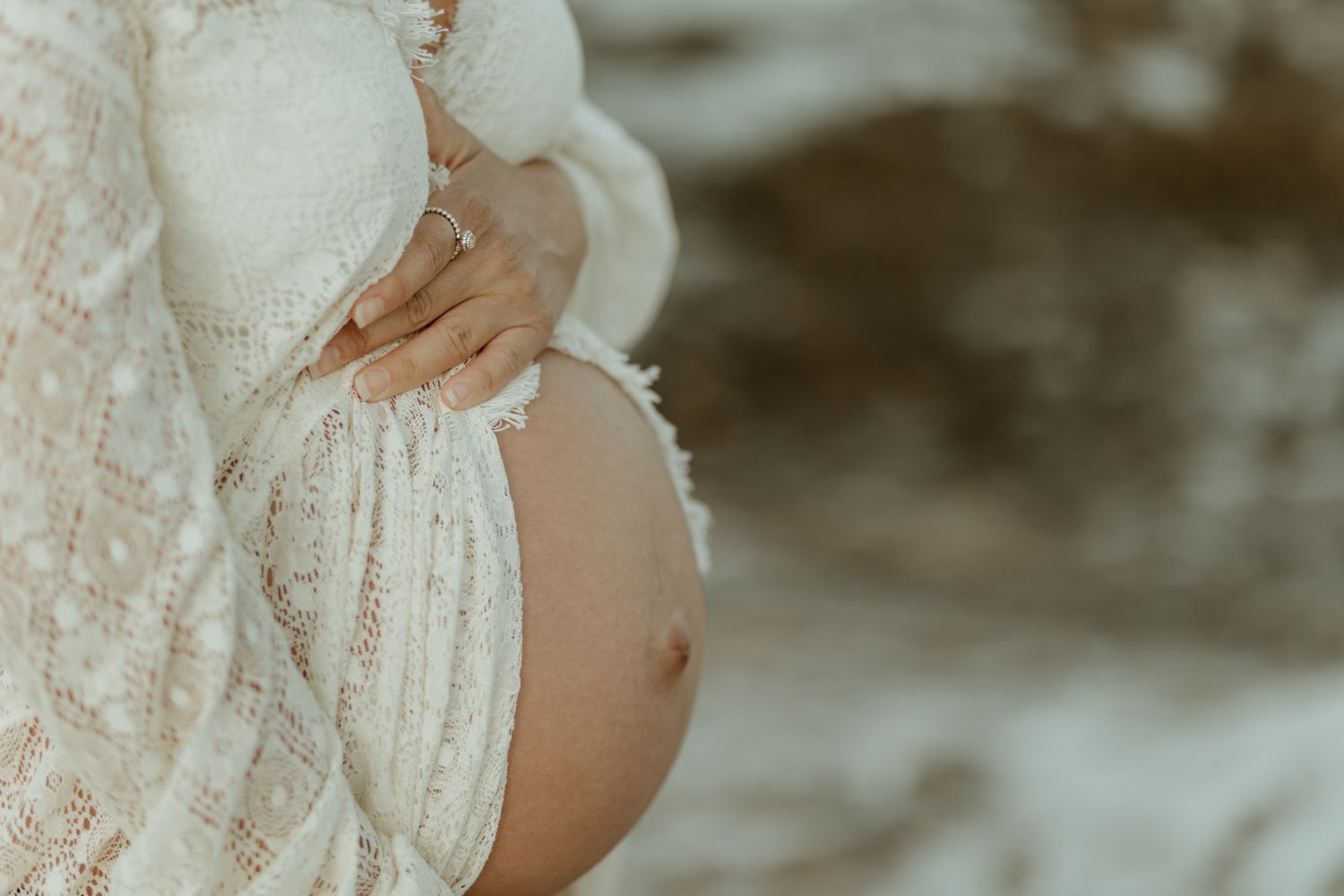 Close-up shot of pregnant belly in white lace dress against ocean backdrop.
