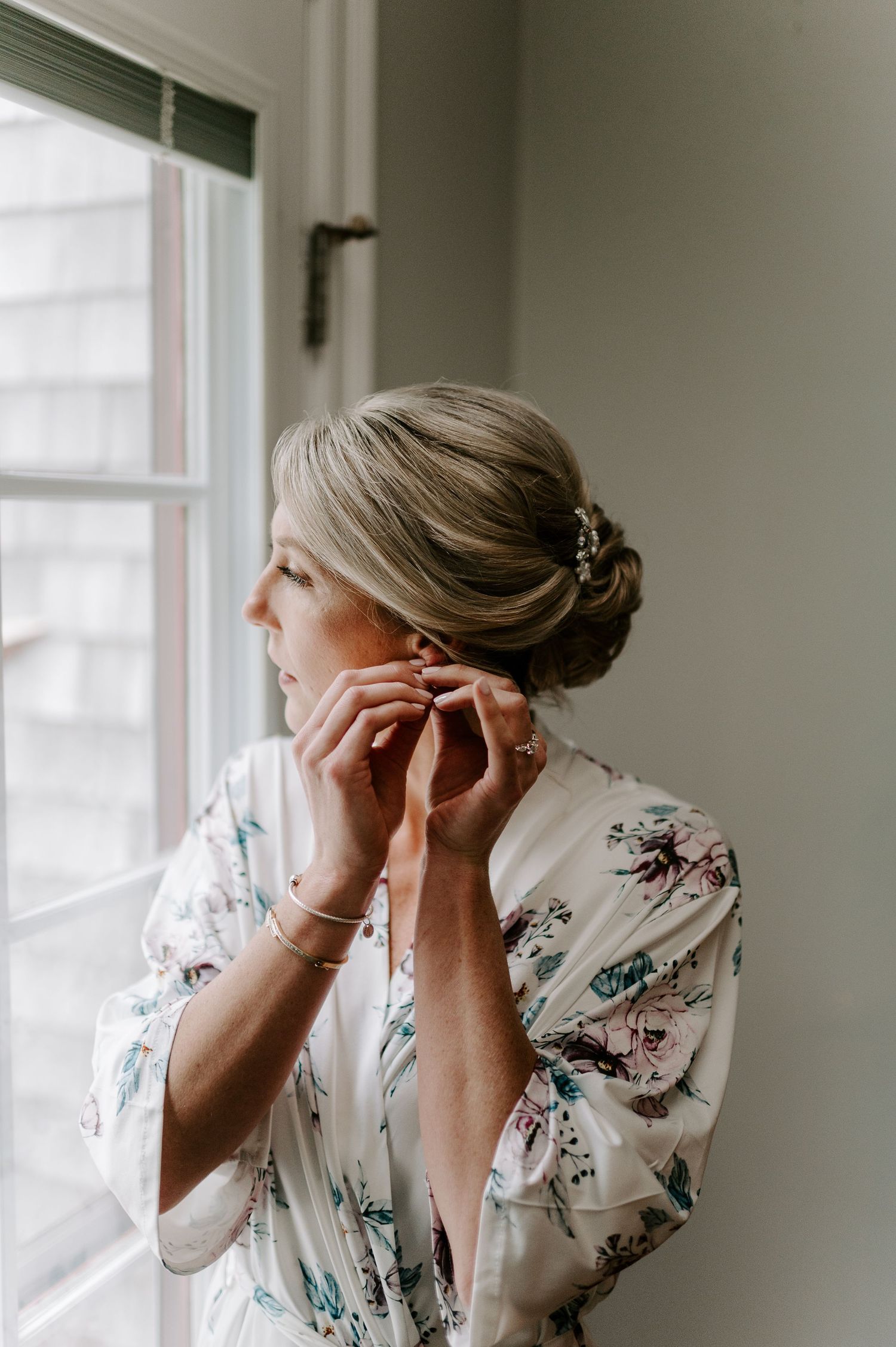 A bride in a floral robe looks out a bright window during wedding preparations.