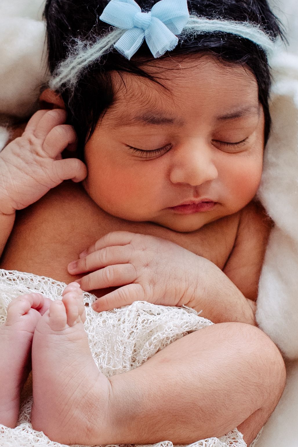 Sleeping newborn baby wearing a light blue hair bow and lace dress rests peacefully on white bedding.
