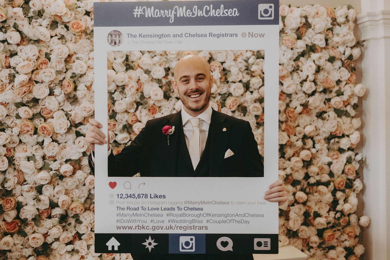 Person holding large social media frame prop against floral backdrop at wedding celebration.
