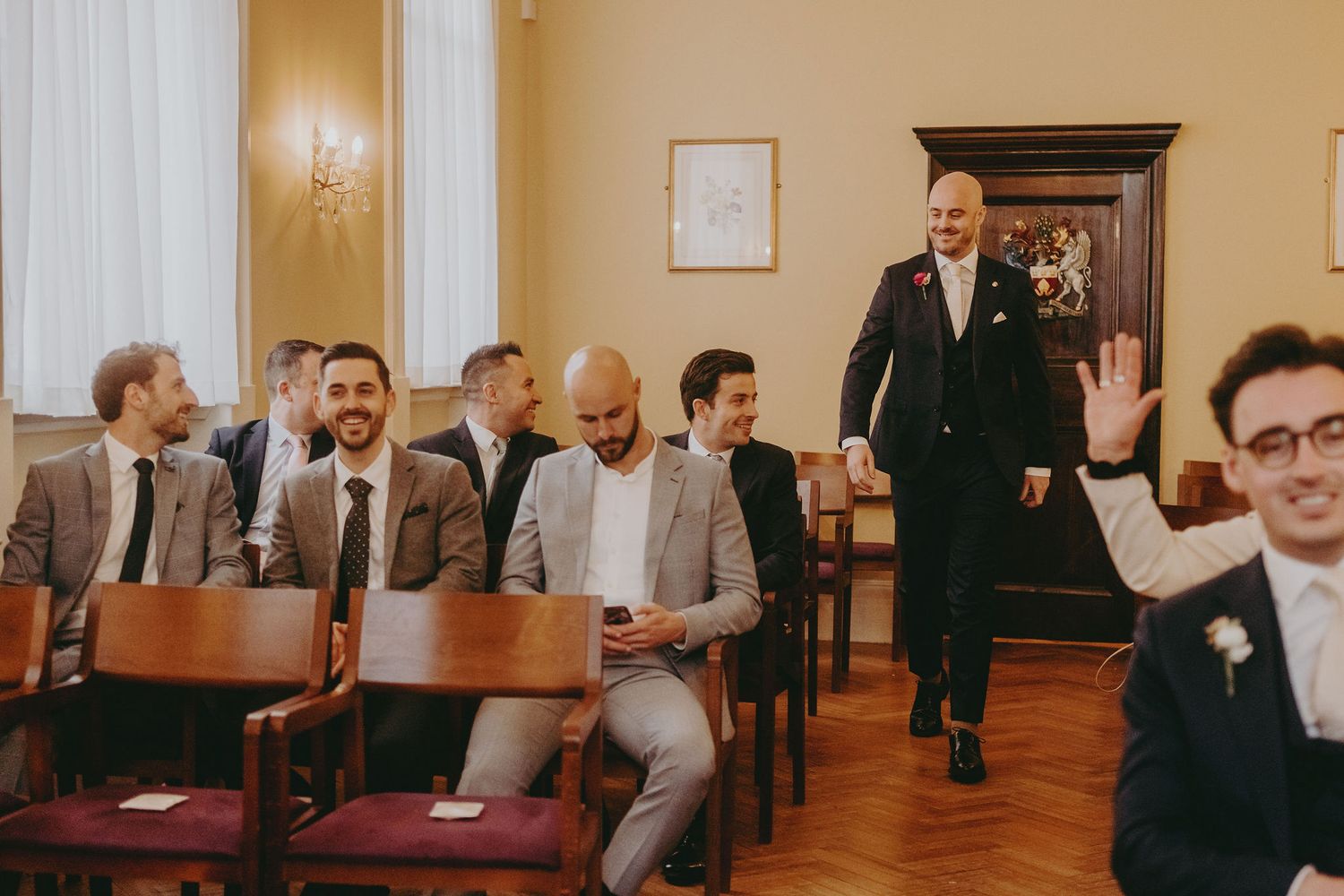 Wedding guests seated in wooden chairs smile and wave in an elegant room with cream walls.