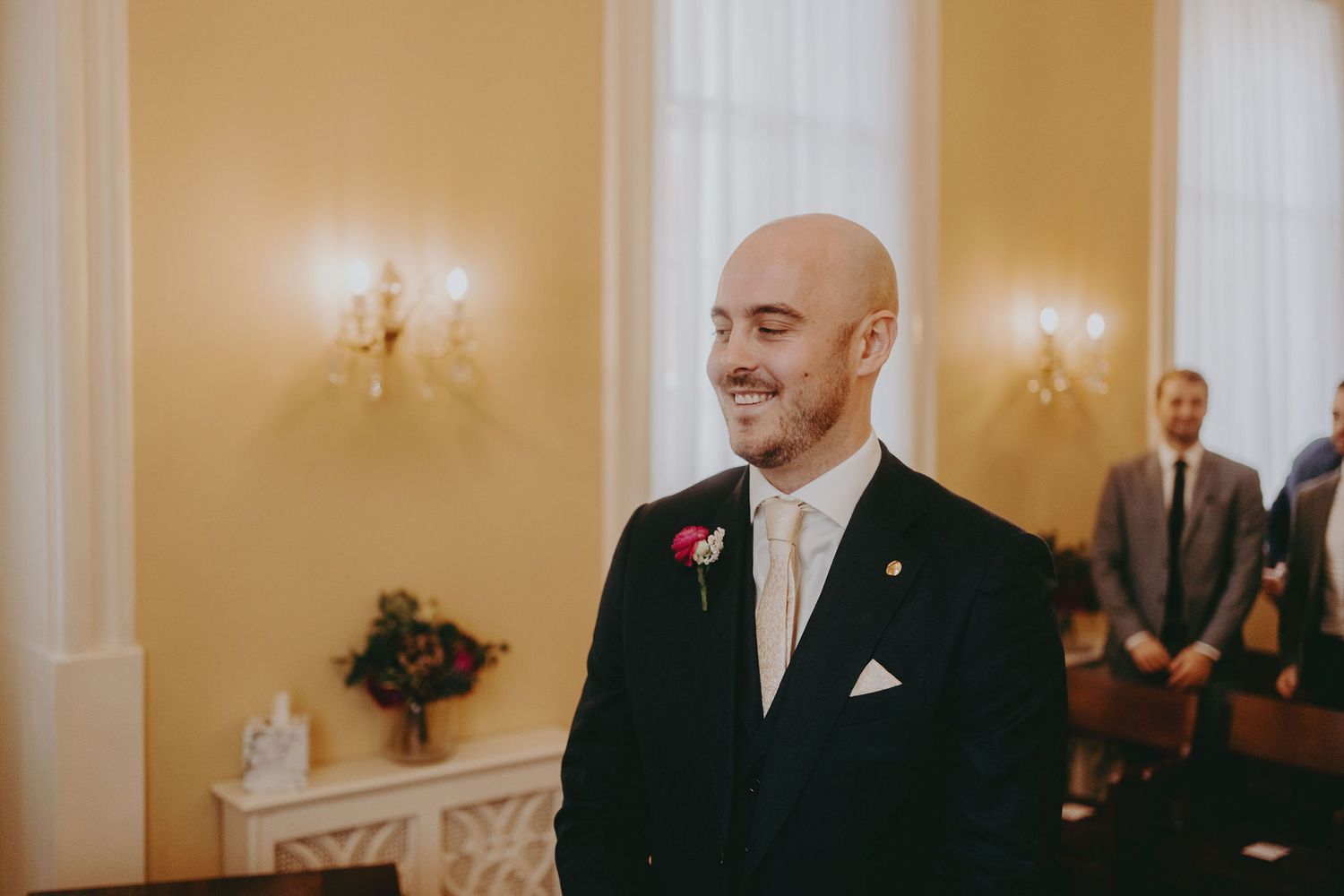 Groom in black suit with white tie stands smiling in elegant room with wall sconces.