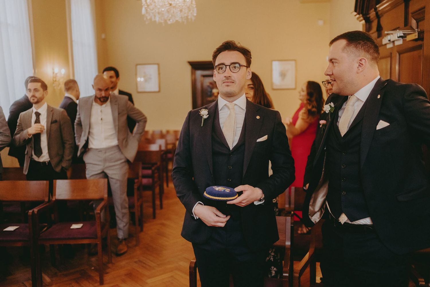 Wedding party members in formal attire gather in room with wooden chairs.