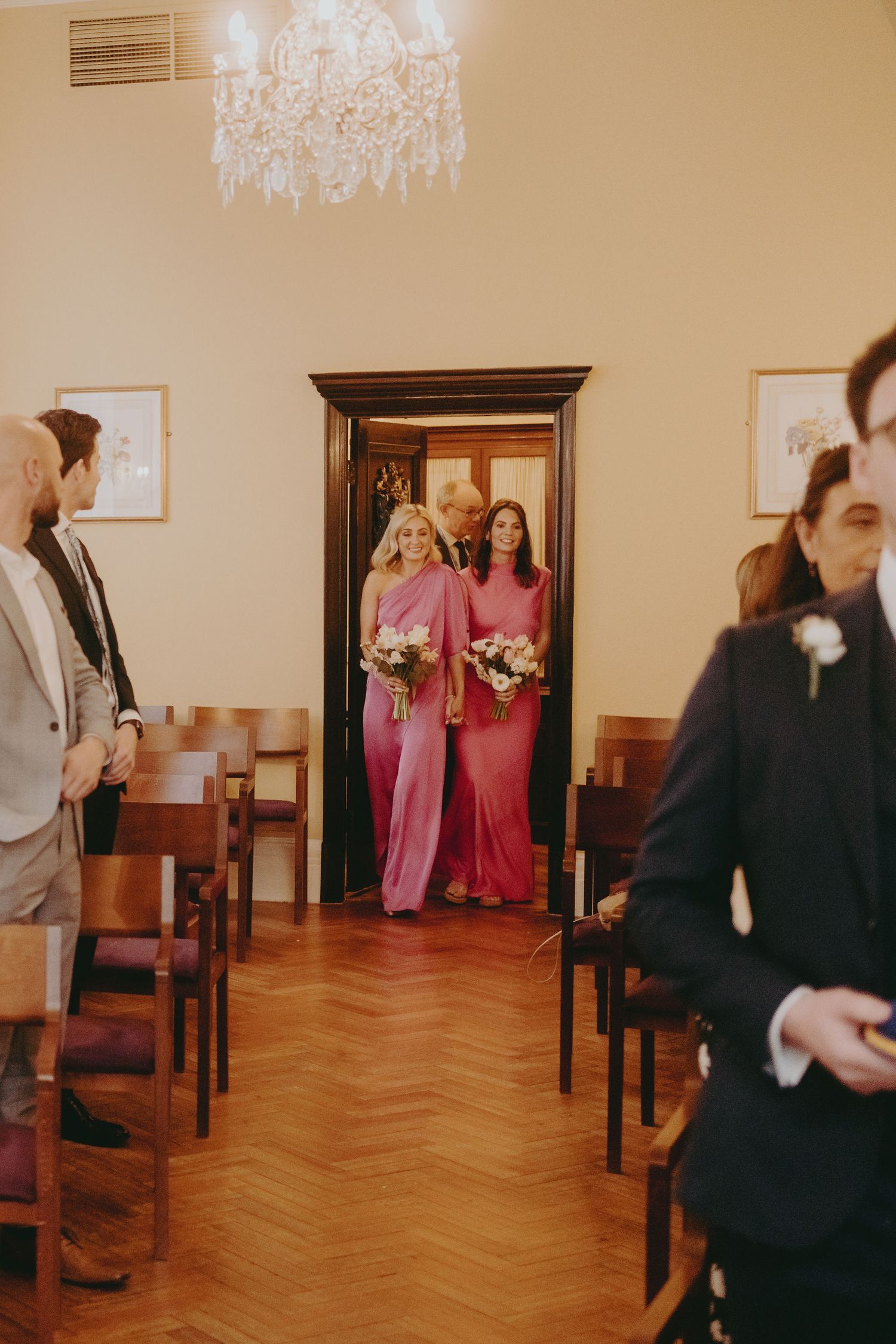 Bridesmaids in pink dresses enter through doorway during wedding ceremony.